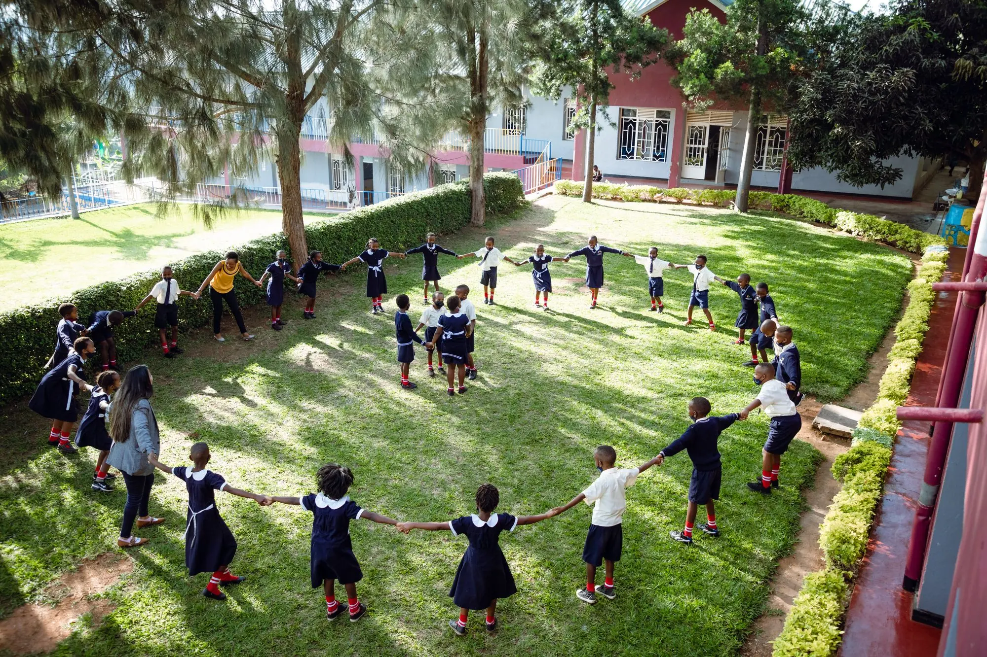 Pupils in navy uniform holding hands in a wide circle on a school lawn, with classroom blocks behind them.