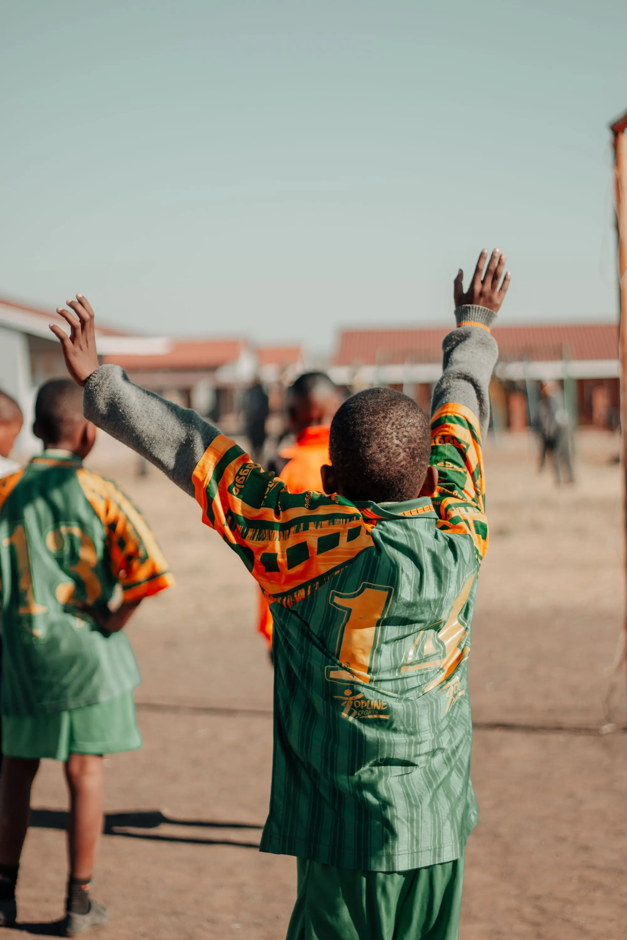 A young footballer with both arms raised, celebrating a goal on a sunny pitch.