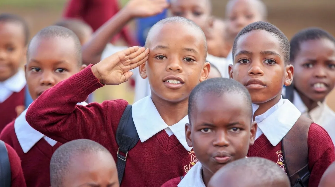 Madaraka pupils in maroon jumpers and white collars standing at morning assembly, one boy saluting during the anthem.
